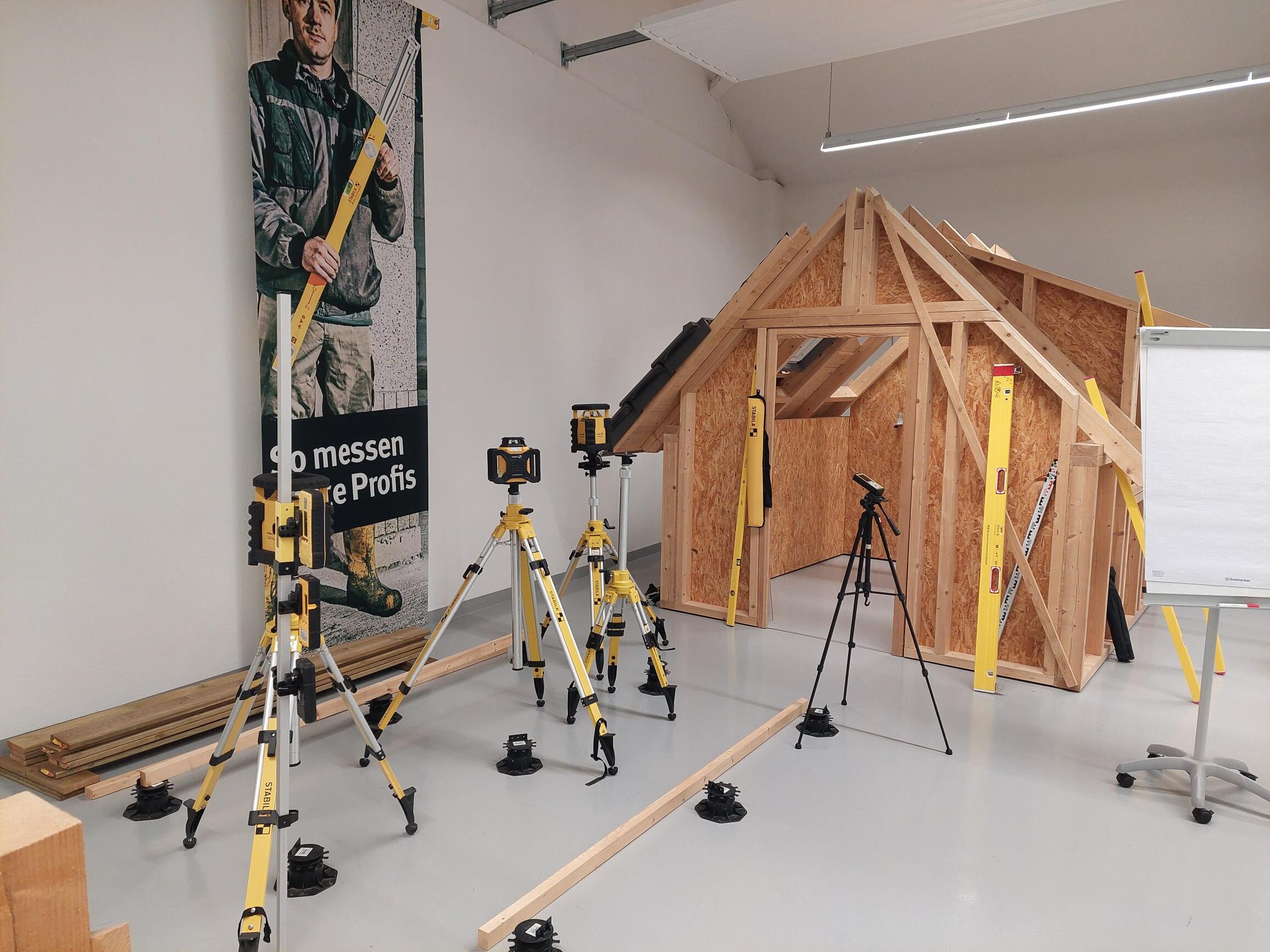 Innenraum mit Holzstrukturhaus, Baugeräten, Stabstativen und Werbebanner, helle Wände.
