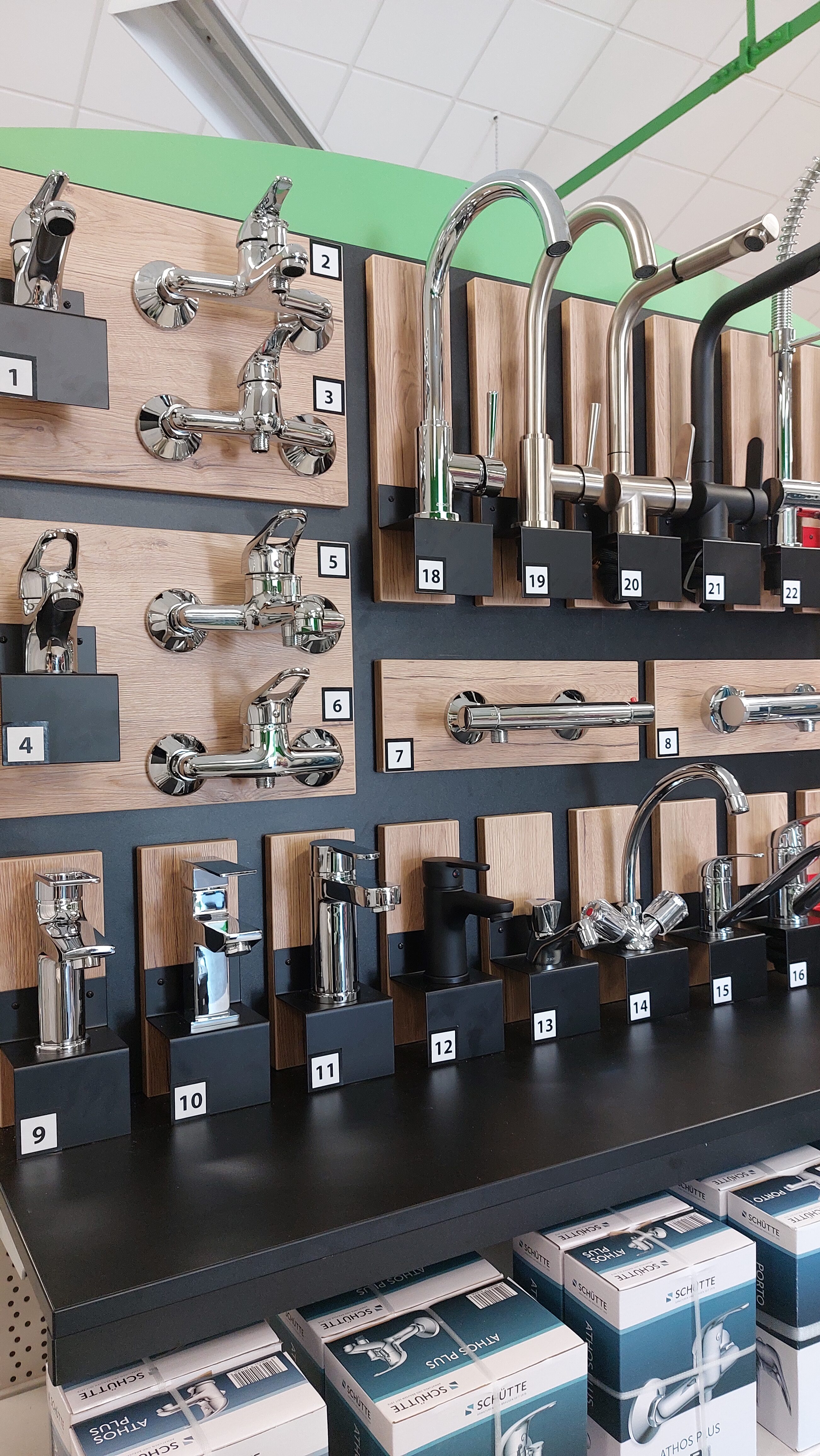 Wand mit verschiedenen Wasserhähnen in Silber auf Holzrückwand in einem Ausstellungsraum.
